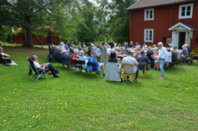 Människor som sitter på bänkar framför röd timrad stuga.