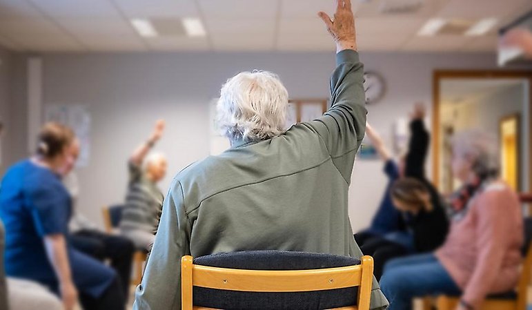 En grupp äldre och personal sitter i ring och sträcker upp en hand i luften.