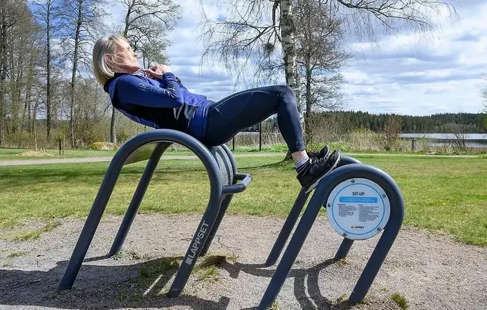 Blond kvinna gör en magövning i en maskin i ett utegym på en gräsmatta.